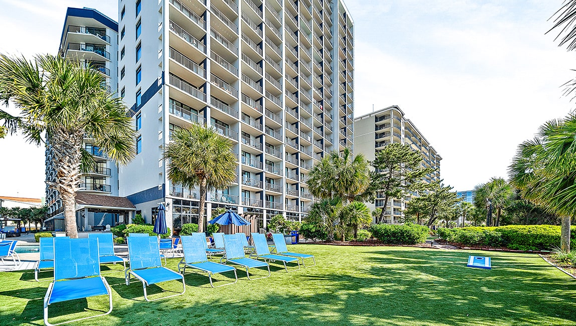 A large white building with many balconies and a pool area with blue chairs and palm trees.
