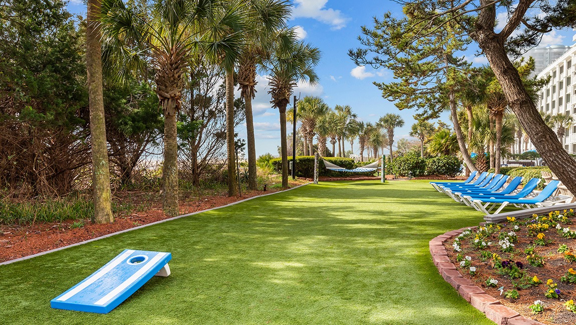A green artificial turf lawn with a blue cornhole game, lounge chairs, and palm trees.